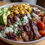 Tender, spice-rubbed steak and smoky charred corn mingle with ripe avocado slices and bright cherry tomatoes over quinoa, finished with silky green cilantro cream sauce and lime wedges.