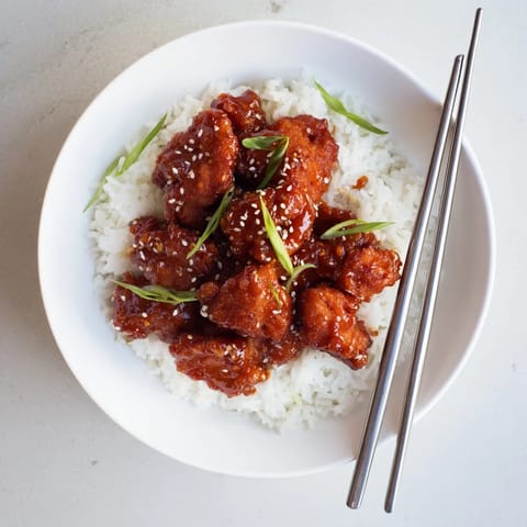 Crispy fried General Tsos Chicken pieces coated in a sticky, tangy sauce with chilies and sesame seeds on a bed of fluffy rice.