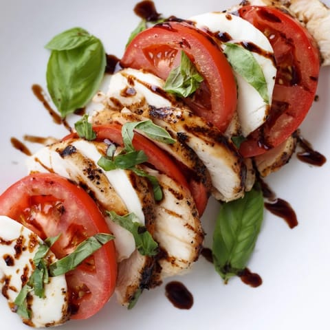 Grilled chicken slices rest atop ripe tomatoes, fresh mozzarella, and basil on a platter, drizzled with tangy balsamic glaze for a vibrant Chicken Caprese Salad.